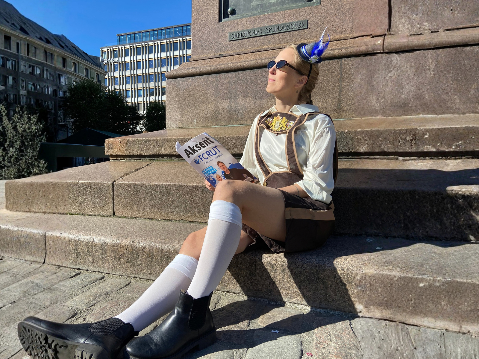 En kvinne kledd i tradisjonelle oktoberfest-klær sitter i solen og leser IOGTs medlemsblad.
